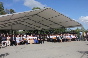 Publiczność siedzi pod dużym białym namiotem podczas dożynkowej uroczystości plenerowej. Wśród uczestników widoczne są osoby w strojach regionalnych, a w tle znajduje się zieleń i błękitne niebo.