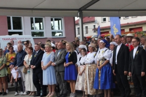 Uczestnicy dożynkowej uroczystości stoją pod zadaszeniem. Wśród nich widoczne są osoby w strojach regionalnych, duchowni oraz mieszkańcy z przypiętymi słonecznikami, a w tle znajdują się budynki i flagi wydarzenia.