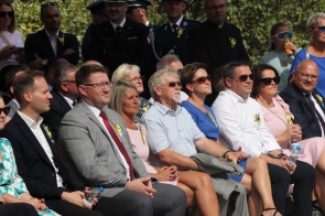 Uczestnicy siedzą wśród publiczności podczas dożynkowej uroczystości plenerowej. Część osób ma przypięte słoneczniki, a w tle widoczni są mundurowi oraz inni obserwujący wydarzenie.