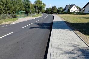 Wyasfaltowana droga biegnie między ogrodzeniami posesji z zielenią i krzewami. W tle widać domy jednorodzinne, a nad nimi niebo z białymi chmurami.