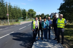 Grupa sześciu osób stoi przy świeżo wyremontowanej drodze asfaltowej z chodnikiem z kostki brukowej. Dwie osoby w odblaskowych kamizelkach, reszta w ubraniach codziennych. W tle widać zieloną wiatę przystankową, ogrodzenie z siatki oraz kilka domów jednorodzinnych otoczonych drzewami. Pogoda jest słoneczna, niebo niemal bezchmurne.