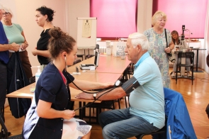 Starszy mężczyzna siedzi przy stole, a pielęgniarka mierzy mu ciśnienie tętnicze za pomocą mankietu i stetoskopu. W tle stoją i rozmawiają inne osoby, a na stole widać medyczne przyrządy.