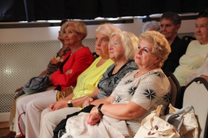 Grupa starszych kobiet siedzi obok siebie w rzędzie, uważnie słuchając tego, co dzieje się przed nimi. Mają spokojne, skupione twarze, a część z nich trzyma torebki na kolanach lub przy krzesłach.