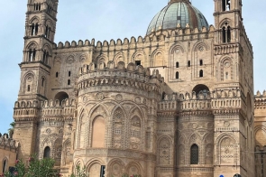 Monumentalna katedra z wielką kopułą i dwiema strzelistymi wieżami, bogato zdobiona arkadami i wzorami kamieniarskimi; przed budowlą niewielki skwer z ławkami i kwitnącymi krzewami, spokojna ulica.