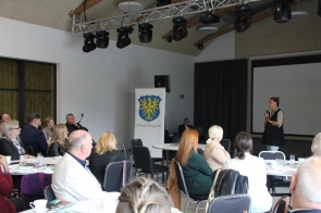 Konferencyjna sala z publicznością przy okrągłych stolikach; po prawej kobieta przemawia z mikrofonem. W tle roll-up z herbem i napisem „Powiat Cieszyński”, nad sceną zawieszone oświetlenie