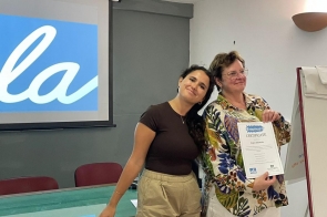 Dwie kobiety w sali szkoleniowej pozują do zdjęcia; jedna opiera głowę na ramieniu drugiej, która trzyma certyfikat Erasmus+. W tle ekran projektora, stół i flipchart.