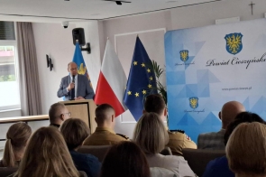 Mówca w garniturze przemawia przy mównicy, trzyma mikrofon. Przed nim siedzi publiczność; w tle stoją flagi Polski, Unii Europejskiej i regionalna oraz ścianka z herbem i napisem „Powiat Cieszyński”.