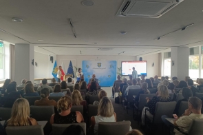 Sala konferencyjna wypełniona uczestnikami; z przodu trzy osoby prowadzą część spotkania przy mównicy i na środku, obok stoi ekran ze slajdem. W tle ścianka z herbem „Powiat Cieszyński” oraz flagi Polski, UE i regionalna.