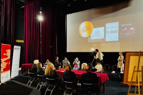 Scena teatru z burgundowymi kotarami, przy długim stole siedzi kilka osób, część fotografuje i nagrywa kamerą. Nad nimi duży ekran z historycznymi grafikami i plakatami teatru. Po bokach stoją roll-upy i sztaluga, w tle rząd krzeseł.