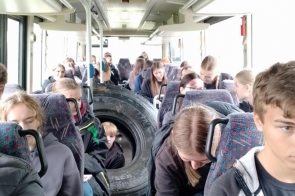 Wnętrze autobusu pełne młodych osób, w przejściu leżą dwa ogromne opony. Kilkoro pasażerów pochyla się lub ma na ciele ślady przypominające urazy, panuje chaos i poruszenie.