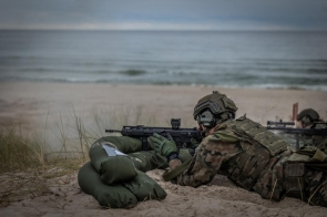 Żołnierz w mundurze maskującym leży na plaży z karabinem snajperskim, opierając broń na workach z piaskiem. W tle widoczne morze i spokojna linia horyzontu.