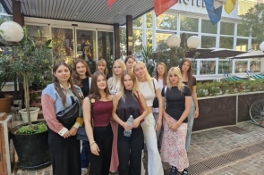 Grupa nastolatek stoi przed wejściem do hotelu pod wiszącymi flagami. Wokół donice z zielenią, za nimi duże okna i napis „HOTEL”. Dziewczyny w codziennych stylizacjach, jedna trzyma butelkę wody, inna plecak.