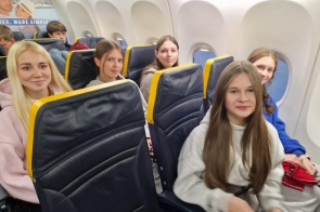 Pięć uśmiechniętych nastolatek siedzi obok siebie w rzędzie w samolocie o żółto-granatowych fotelach. Za oknami jasne światło, w tle widać innych pasażerów.