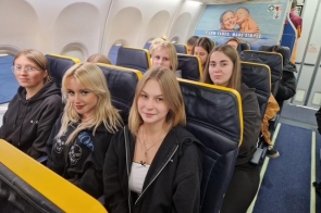 Siedem nastolatek w samolocie, w dwóch rzędach żółto-granatowych foteli, większość w ciemnych, wygodnych bluzach, uśmiechnięte. Po prawej wąskie przejście, nad głowami jasne oświetlenie. Na ścianie z tyłu reklama z dwojgiem bawiących się niemowląt.