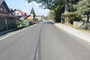Asfaltowa ulica biegnąca przez miejscowość z domami po lewej stronie i drzewami po prawej. Na jezdni widoczne są linie podziału, a w tle widać zaparkowane samochody i fragment nieba.