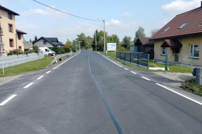 Nowa asfaltowa droga biegnąca przez miejscowość z domami po obu stronach. Na jezdni widoczne są białe linie krawędziowe. W tle widać ogrodzenia, zieleń i fragment nieba.
