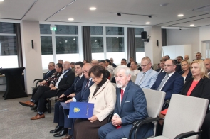 Grupa osób siedzi w rzędach w sali sesyjnej. Większość ma na sobie formalne stroje, a kilka osób trzyma dokumenty i teczki. Uczestnicy są zwróceni w stronę niewidocznej części sali.