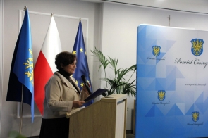 Janina Żagan Starosta Cieszyński przemawia przy mównicy, trzymając mikrofon oraz teczkę. W tle widoczne są flagi oraz kawałek ścianki Powiatu Cieszyńskiego.