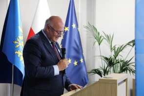 Jan Kawulok Dyrektor Cieszyńskiego Pogotowia Ratunkowego przemawia przy mówicy, trzymając mikrofon. W tle widoczne są flagi oraz roślina doniczkowa.