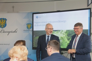 Mieczysław Szczurek Wicestarosta Cieszyński oraz Jerzy Pilch Członek Zarządu stoją przed ekranem z prezentacją. W tle widoczna jest ścianka z herbem Powiatu Cieszyńskiego, a na pierwszym planie siedzą uczestnicy spotkania.