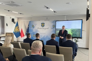 Mężczyzna prowadzi prezentację w sali konferencyjnej przed siedzącymi uczestnikami. Na ekranie widoczny jest slajd dotyczący programu redukcji ryzyka powodziowego.