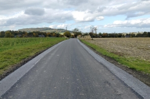 Wyremontowana droga asfaltowa. Po obu stronach widoczne są utwardzone pobocza, a w tle znajdują się pojedyncze zabudowania, drzewa oraz pagórkowaty krajobraz pod zachmurzonym niebem.