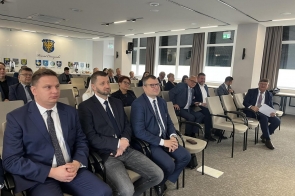 Na pierwszym planie widoczni są mężczyźni w garniturach, a w tle pozostali uczestnicy spotkania, herb powiatu oraz herby gmin.