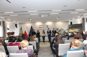 Uczestnicy siedzą, zwróceni w stronę występującej młodzieży. Przy mikrofonie stoją uczniowie, a w tle widoczne są flagi, godło Polski, herb Powiatu oraz ekran z prezentacją.