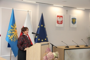 Janina Żagan Starosta Cieszyński przemawia przy mównicy. W tle widoczne są flagi, godło Polski, herb Powiatu Cieszyńskiego oraz stół prezydialny z mikrofonami.