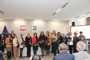Grupa nagrodzonych osób pozuje. Nagrodzeni trzymają róże i teczki, a w tle widoczne są godło Polski, herb Powiatu Cieszyńskiego, flaga Unii Europejskiej oraz ekran z grafiką wydarzenia.