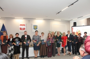 Grupa nagrodzonych osób pozuje. Nagrodzeni trzymają róże i teczki, a w tle widoczne są godło Polski, herb Powiatu Cieszyńskiego, flaga Unii Europejskiej oraz ekran z grafiką wydarzenia.