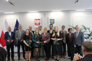 Grupa nagrodzonych osób pozuje. Nagrodzeni trzymają róże i teczki, a w tle widoczne są godło Polski, herb Powiatu Cieszyńskiego oraz flaga Unii Europejskiej.