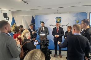 Konferencja prasowa w sali ze ścianką Powiatu Cieszyńskiego. Przedstawiciele władz stoją przed dziennikarzami, mikrofonami i kamerami, a w tle widoczne są flagi.