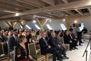 Duża grupa ludzi siedzi w jasnej sali z drewnianymi belkami na suficie, uważnie słuchając osoby przemawiającej poza kadrem. Większość uczestników jest elegancko ubrana — mężczyźni w garniturach, kobiety w formalnych strojach. Pomieszczenie jest dobrze oświetlone, z oknami dachowymi i reflektorami skierowanymi na scenę.