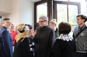Grupa osób rozmawia w jasnym pomieszczeniu przy dużych przeszklonych drzwiach. Ludzie są elegancko ubrani i uśmiechnięci. Mężczyzna w okularach i ciemnej kurtce gestykuluje podczas rozmowy z kobietą w czarnej sukni z białym koronkowym kołnierzem oraz inną kobietą w ozdobnej opasce z piórami. W tle widać pozostałych uczestników spotkania oraz widok na zieleń za oknem.
