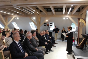 W przestronnej sali z drewnianymi belkami na suficie, Janina Żagan Starosta Cieszyński w czarnej sukni z białym koronkowym kołnierzem przemawia do siedzącej publiczności. Trzyma mikrofon i kartkę z notatkami. Przed nią siedzą elegancko ubrani goście w garniturach i formalnych strojach, uważnie słuchając. W tle widać harfę, sprzęt oświetleniowy i osoby stojące przy ścianie.
