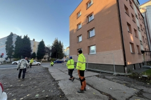 Pracownicy w odzieży odblaskowej znajdują się przy budynku, obok którego prowadzone są prace remontowe. Widać fragment ogrodzenia zabezpieczającego teren, nierówną nawierzchnię oraz sąsiednią ulicę.