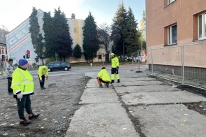 Pracownicy w odzieży odblaskowej prowadzą prace przy budynku. Widoczne są płyty betonowe, ogrodzenie zabezpieczające teren remontu oraz ulica i zabudowania w tle.