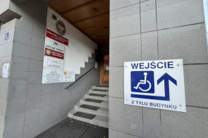 Wejście do Powiatowego Urzędu Pracy w Cieszynie. Przy schodach widoczne są tablice informacyjne urzędu, a po prawej stronie znajduje się oznaczenie wskazujące wejście dla osób z niepełnosprawnościami z tyłu budynku.
