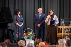 Jubileusz LO im. Pawła Stalmacha. Na zdjęciu gratulacje składają Przewodniczacy Rady Powiatu Stanisław Kubicius i Janina Żagan Starosta Cieszyński, a obok widoczny jest kosz z kwiatami, fortepian, dekoracje z wrzosów oraz siedząca publiczność.