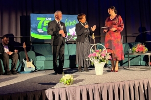75-lecie ZS im. W. Szybińskiego moment wręczenia laudcji przez samorząd powiatowy. Trzy osoby stoją na scenie podczas uroczystości jubileuszowej. Dwie osoby trzymają mikrofony, a jedna dokumenty. W tle widoczny jest ekran, zielona kanapa, gitara oraz dekoracje kwiatowe.