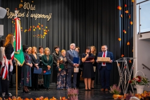 Grupa osób stoi na scenie podczas jubileuszu. Jedna z osób przemawia do mikrofonu, obok mężczyzna trzyma ozdobne pudełko, a w tle widoczny jest napis „80 lat. Przystań i wspomnij”, jesienne dekoracje oraz poczet sztandarowy.