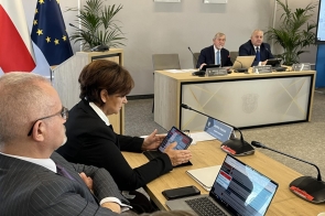 Radni siedzą w sali sesyjnej podczas obrad. Na stołach widoczne są mikrofony, laptop, tablet i dokumenty, a w tle znajdują się flagi, rośliny oraz stanowisko prezydialne.