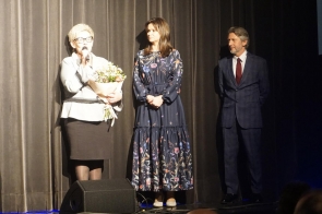 Kobieta w jasnym żakiecie i ciemnej spódnicy trzyma mikrofon oraz bukiet kwiatów, stojąc na scenie przed ciemną kotarą; obok niej kobieta w długiej, granatowej sukni w kwiaty i mężczyzna w eleganckim garniturze patrzą z uwagą w jej stronę