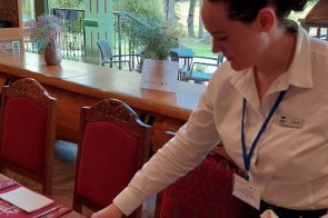 Młoda kobieta w eleganckiej białej koszuli nakrywa do stołu w restauracji o górskim, drewnianym wystroju. Starannie układa sztućce i serwetki na czerwonych podkładkach z motywem reniferów. Przez duże okna widać zielone drzewa i taras.
