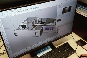 Sala komputerowa, w której kilka osób pracuje przy biurkach z monitorami. Na pierwszym planie ekran z programem do projektowania 3D pokazuje model wnętrza budynku. Na biurku leży kartka, a w tle uczestnicy skupieni na pracy.
