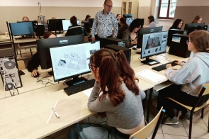 Sala komputerowa, w której uczniowie pracują przy biurkach z komputerami. Na monitorach widać program do projektowania 3D z modelami wnętrz. Wśród pracujących osób przechadza się nauczyciel, obserwując postępy.