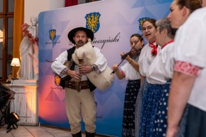Osoby w strojach regionalnych występują na tle ścianki z herbami Powiatu Cieszyńskiego. Mężczyzna trzyma tradycyjny instrument, a obok stoją kobiety śpiewające i grające na skrzypcach.