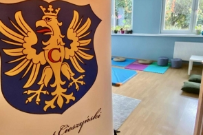 Baner z herbem Powiat Cieszyński stoi przy wejściu do jasnej sali z drewnianą podłogą. W środku widać kolorowe maty, poduszki i kilka roślin ustawionych na parapecie pod dużymi oknami.