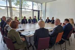 Osoby siedzą przy dużym stole w jasnej sali konferencyjnej. Na blacie widoczne są dokumenty, długopisy i szklanki, a w tle znajdują się duże okna z widokiem na zieleń.